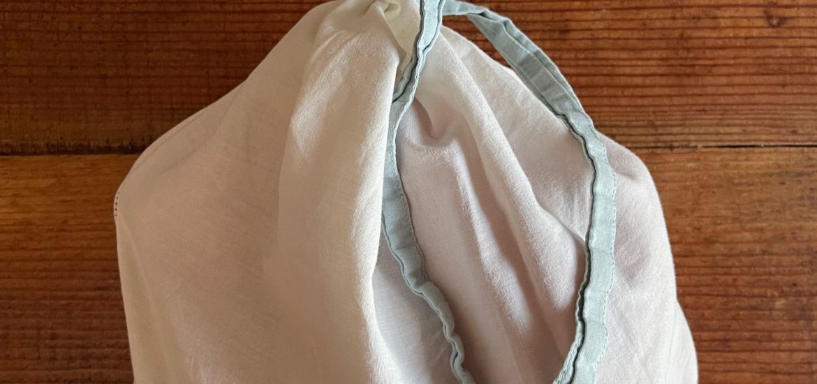 A filled white cloth produce bag is closed at the top with a blue tie. It sits on a dark wooden background.