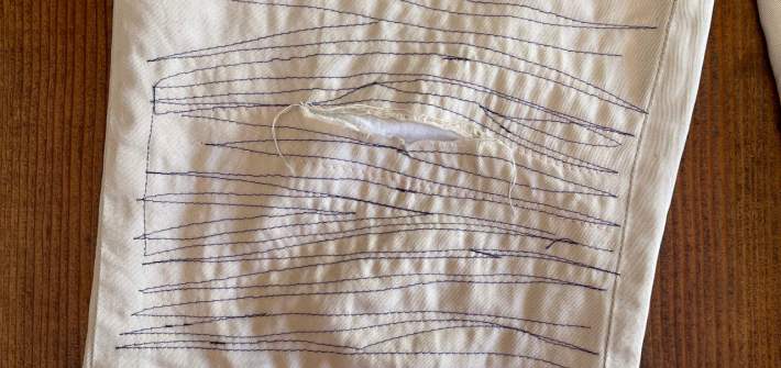 The right pant leg of a beige pair of pants mended with a white patch in the interior. Dark blue thread has been stitched back and forth across the leg around the knee.
